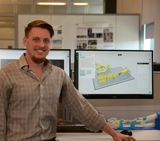 Engineer smiling at a workstation with dual monitors displaying a 3D anatomical model being prepared for printing, with printed medical model parts on the desk.