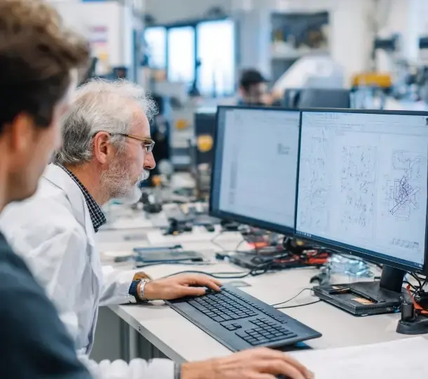 Engineers in a lab review detailed technical drawings and CAD models for a medical device on dual computer monitors, surrounded by electronic equipment and prototype components.