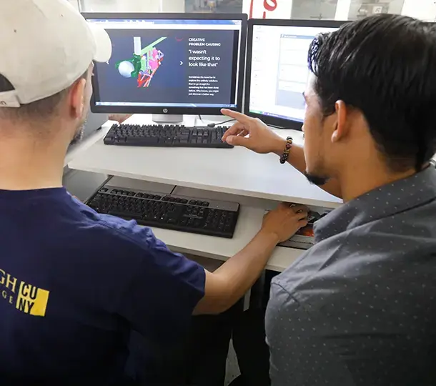Two students working together at a computer workstation, reviewing a 3D model and technical instructions on dual monitors during a design or engineering training session.