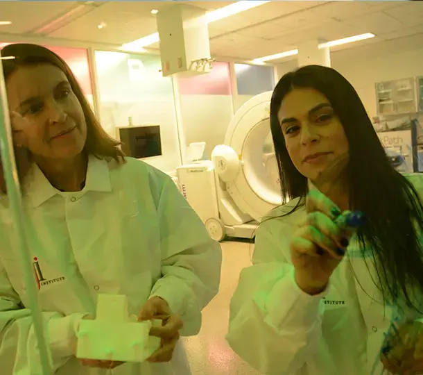 Two clinicians in lab coats reviewing a 3D printed medical device part in a hospital innovation lab, with imaging equipment visible in the background as one of them writes on a glass board.