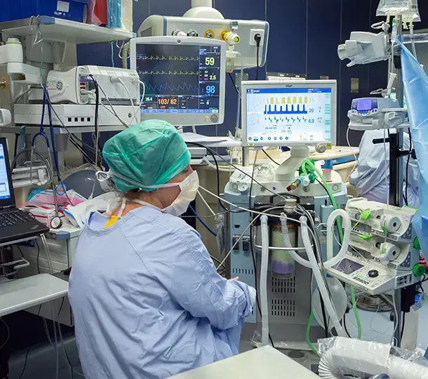 Medical staff in full protective attire operating monitoring and life-support equipment in an intensive care unit, surrounded by ventilators, patient monitors, and infusion devices.