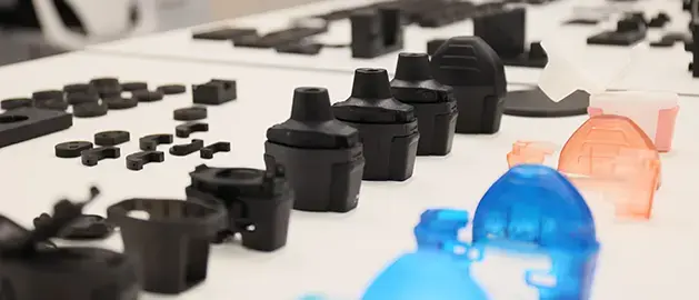 Assorted 3D printed medical device prototype components, including black medical device housings and translucent blue and orange parts, arranged on a white tabletop for evaluation.