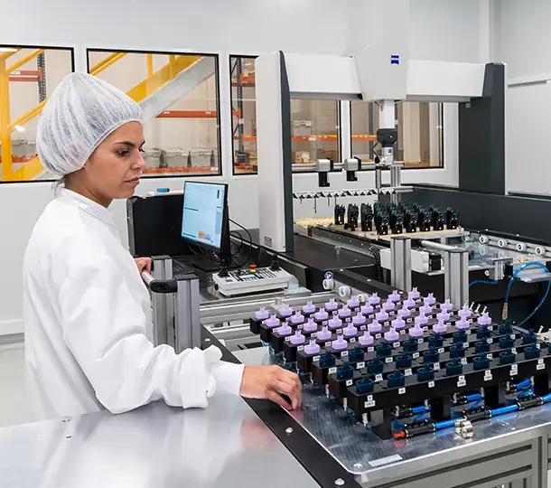 Technician in cleanroom attire operating quality-control equipment on a production line, inspecting rows of assembled medical device components beneath an automated measurement system.