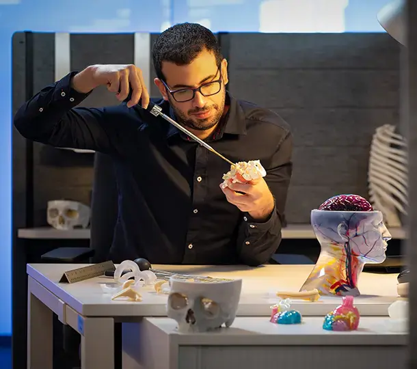 Tech using a tool to examine a 3D-printed anatomical model, surrounded by various medical 3D-printed parts including skulls, organs, and a transparent head model with a visible brain.