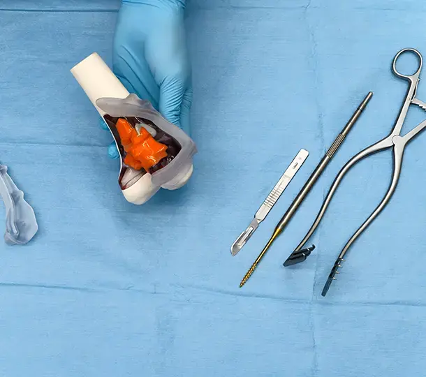 A gloved hand holds a 3D printed anatomical knee model beside various surgical instruments laid out on a blue sterile drape